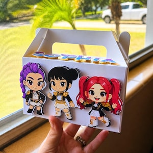 Könnte beinhalten: Eine weiße Giebelbox, verziert mit drei Aufklebern von Zeichentrickfiguren. Die Aufkleber zeigen Mädchen mit lila, schwarzen und roten Haaren, die schwarze und goldene Outfits tragen. Auf der Box steht der Name "Sophia" in blauen und rosa Buchstaben.