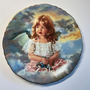 May include: A decorative plate featuring a young girl with red hair, blue eyes, and angel wings. She wears a white lace top and a pink dress, seated on a cloud. The plate has a gold rim and a painted sky background.