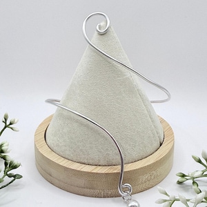 Peut inclure: Un bracelet de bras argenté au design délicat. Le bracelet présente un détail en spirale en haut et un petit charme de perle rond. Le bijou est présenté sur un présentoir conique de couleur claire.
