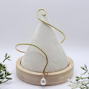 May include: A gold-colored arm cuff with a spiral design at the top and a teardrop-shaped pearl pendant. The cuff is displayed on a white fabric-covered cone, set on a wooden base, with white flowers in the background.
