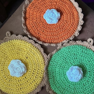 May include: Three circular crochet pot holders in orange, yellow, and green, each featuring a central white crocheted flower and a scalloped tan border. Each piece includes a small loop at the top edge for hanging, showcasing a textured yarn finish.