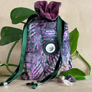 Op de afbeelding: Een paars en groen batikstoffen trekkoordtasje met een donkergroen fluwelen lintsluiting. De tas heeft een grote witte knoop met een decoratief ontwerp. De tas is omgeven door groene bladeren en kleine kristallen.