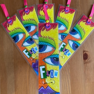 May include: A collection of colorful, artistic bookmarks. Each bookmark features a stylized face with large blue eyes, yellow hair, and the word "FLOW". They have pink ribbon attachments.