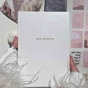 Op de afbeelding: Een wit notitieboek met de woorden "DIG DEEPER" in gouden letters. Het notitieboek wordt vastgehouden door een persoon, omringd door kaarten en decoratieve elementen. Het notitieboek is een hardcover boek.