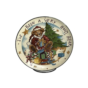 Puede incluir: Un plato de Navidad de cerámica blanca con una ilustración verde, roja y marrón de un oso de peluche con un gorro de Papá Noel sentado sobre una pila de regalos. El texto "I've Been A Very Good Bear" está escrito alrededor del borde del plato.