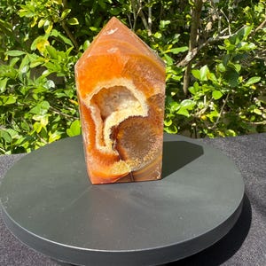 Könnte beinhalten: Ein polierter orangefarbener Achat-Kristallturm mit spitzer Spitze. Der Kristall hat eine ausgehöhlte Mitte mit einem weißen und gelben Inneren. Er steht auf einem schwarzen, rotierenden Präsentationsständer.