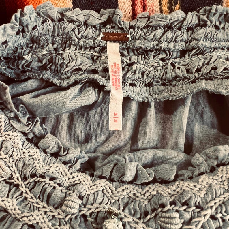 May include: Close-up of a light blue top with intricate crochet and ruffled details. The top features a shirred neckline and a tag with the brand name and size. The fabric appears soft and lightweight, suitable for warm weather.