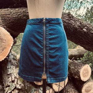 Pu&ograve; includere: Una gonna di jeans blu con una cerniera argentata sul davanti. La gonna &egrave; esposta su un manichino. Lo sfondo presenta tronchi e rami di alberi.