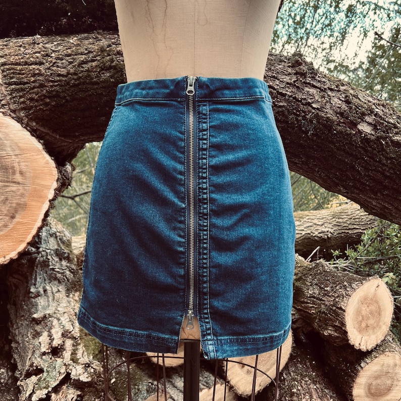 Pu&ograve; includere: Una gonna di jeans blu con una cerniera argentata sul davanti. La gonna &egrave; esposta su un manichino. Lo sfondo presenta tronchi e rami di alberi.