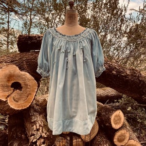 May include: A light blue dress with short sleeves and a round neckline. The dress features delicate embroidery around the neckline and cuffs, with small decorative buttons down the front. The dress is displayed on a mannequin.