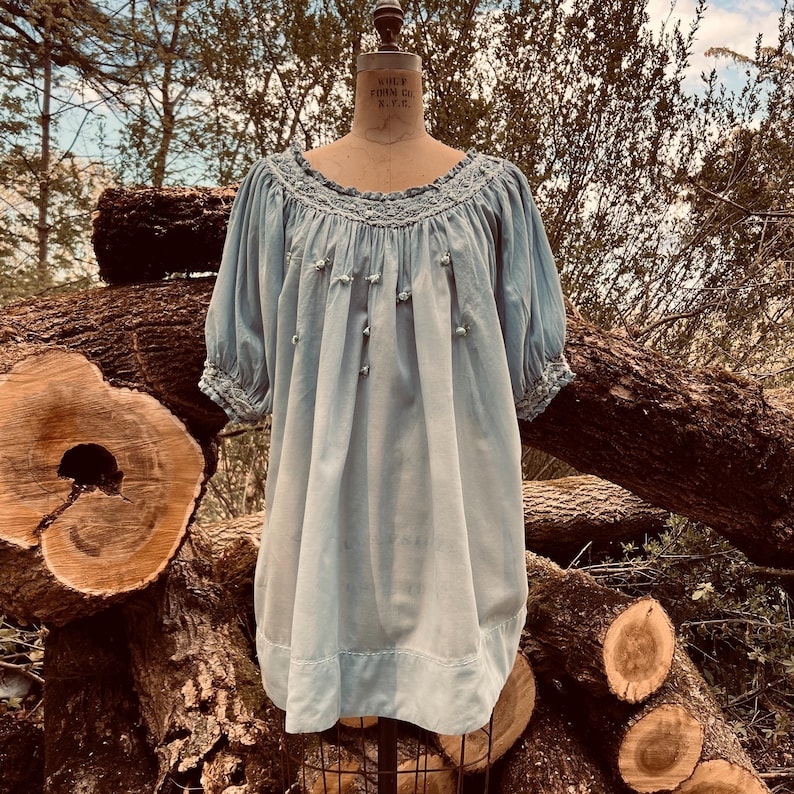May include: A light blue dress with short sleeves and a round neckline. The dress features delicate embroidery around the neckline and cuffs, with small decorative buttons down the front. The dress is displayed on a mannequin.
