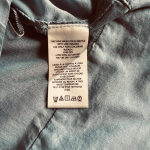 May include: A close-up of a clothing tag with care instructions in English and French. The tag is white with black text and is sewn onto a light blue fabric. The tag includes washing, drying, and ironing instructions.