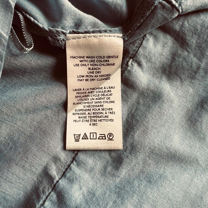 May include: A close-up of a clothing tag with care instructions in English and French. The tag is white with black text and is sewn onto a light blue fabric. The tag includes washing, drying, and ironing instructions.