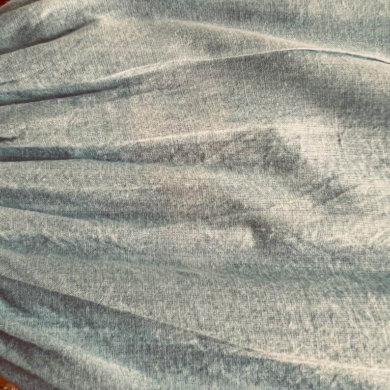 May include: Close-up of a textured, light blue fabric with soft folds and subtle variations in color. The fabric appears to be lightweight, possibly linen or a similar material, with a slightly wrinkled appearance.