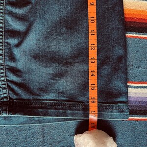 Pu&ograve; includere: Primo piano di tessuto denim blu scuro con un metro a nastro arancione. Il nastro mostra una misura di circa 23-46 cm. Un cristallo bianco &egrave; in basso nell'immagine.