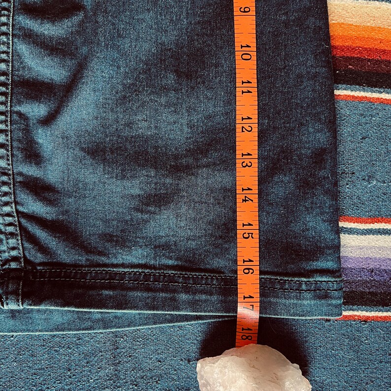 Pu&ograve; includere: Primo piano di tessuto denim blu scuro con un metro a nastro arancione. Il nastro mostra una misura di circa 23-46 cm. Un cristallo bianco &egrave; in basso nell'immagine.