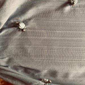May include: A close-up of a light blue fabric with a subtle striped texture. The fabric is adorned with several small, decorative floral embellishments. The embellishments appear to be made of a light-colored material, possibly fabric or a similar textile.