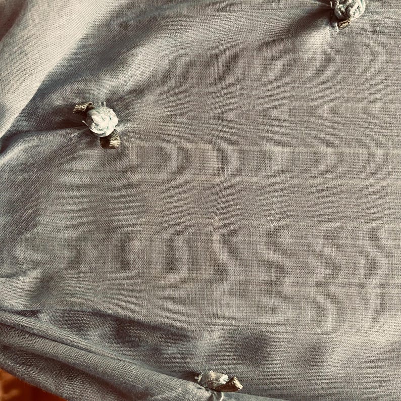 May include: A close-up of a light blue fabric with a subtle striped texture. The fabric is adorned with several small, decorative floral embellishments. The embellishments appear to be made of a light-colored material, possibly fabric or a similar textile.