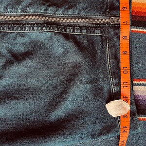 Pu&ograve; includere: Primo piano di tessuto denim blu scuro con cerniera argentata. Un metro a nastro arancione mostra una lunghezza di circa 17.8 cm. &Egrave; presente anche un piccolo cristallo trasparente di forma irregolare.