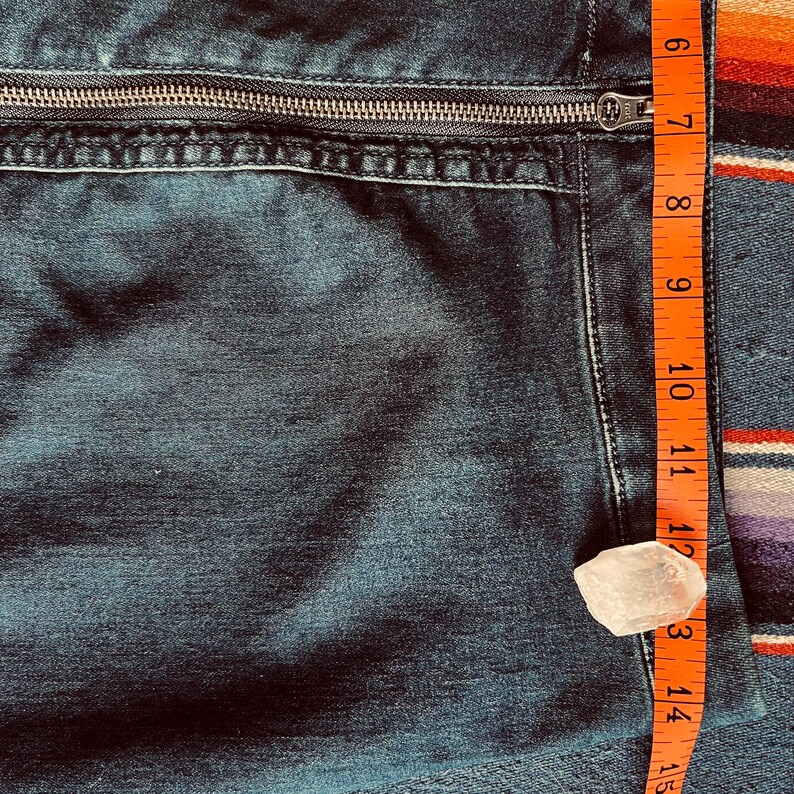 Pu&ograve; includere: Primo piano di tessuto denim blu scuro con cerniera argentata. Un metro a nastro arancione mostra una lunghezza di circa 17.8 cm. &Egrave; presente anche un piccolo cristallo trasparente di forma irregolare.