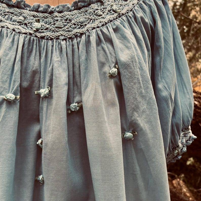 May include: A light blue blouse with a smocked neckline and short sleeves. The blouse features small, decorative rosettes and a gathered design. The fabric has a soft, flowing appearance, suggesting a lightweight material.