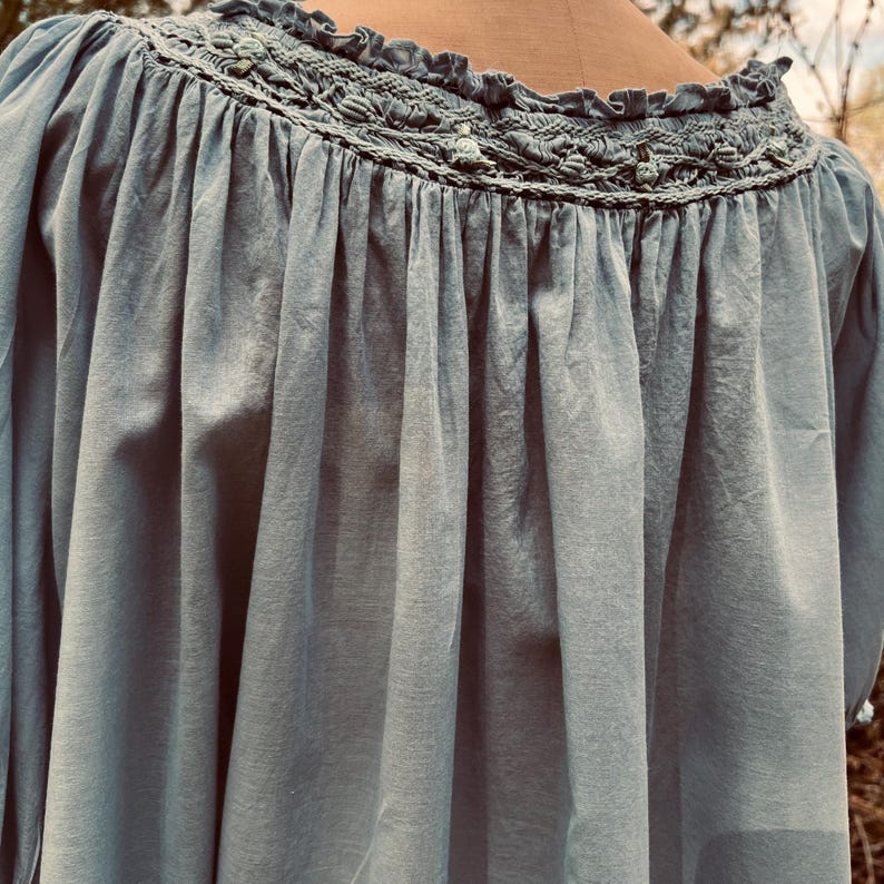 May include: A light blue long-sleeve top with a detailed, embroidered neckline. The top has a loose, flowing fit and gathered fabric below the neckline. The neckline features a decorative, textured design with small, light green accents.