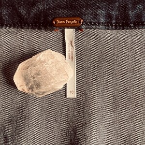 Pu&ograve; includere: Primo piano di un tessuto denim scuro con un'etichetta Free People. Un cristallo rosa chiaro &egrave; appoggiato sul tessuto vicino a un'etichetta che recita "Made in China, Fabriqu&eacute; en Chine" e il numero 10.