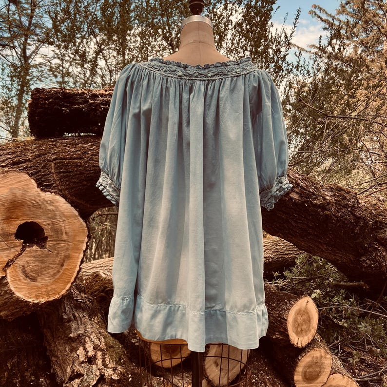 May include: A light blue peasant top with a ruffled neckline and sleeves. The top is displayed on a mannequin against a backdrop of tree trunks and foliage. The top has a relaxed fit and a soft, flowing appearance.