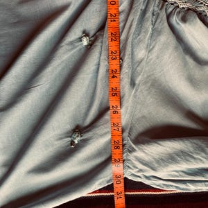 May include: A light blue garment with decorative knot details. An orange measuring tape shows the length of the garment to be approximately 30 inches. The fabric has a soft, flowing appearance.