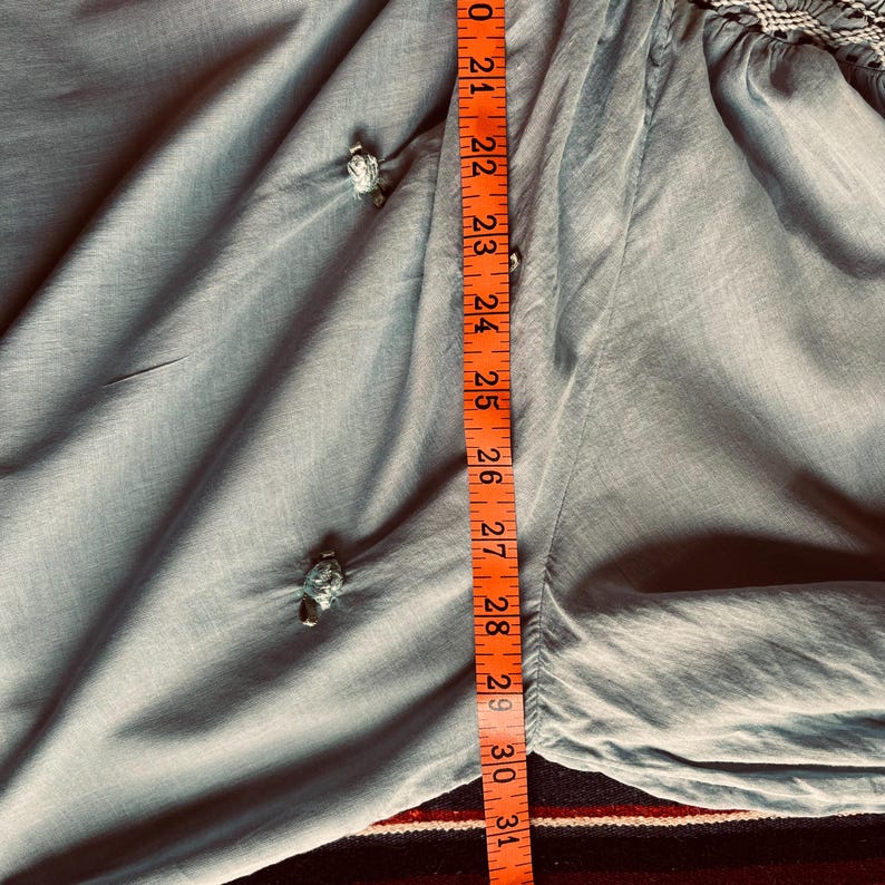 May include: A light blue garment with decorative knot details. An orange measuring tape shows the length of the garment to be approximately 30 inches. The fabric has a soft, flowing appearance.