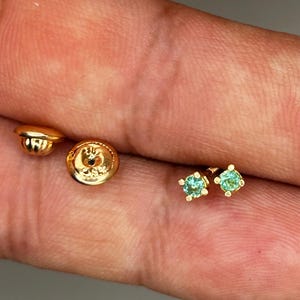 May include: Close-up of gold-colored earrings with small, round, light blue gemstones. The earrings are set in a four-prong setting. Also visible are two gold earring backs. Jewelry.