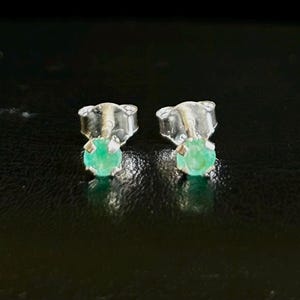 May include: A pair of stud earrings featuring round, vibrant green gemstones set in silver-colored metal. The earrings are displayed against a dark, textured background, highlighting the gemstones' color and shine.