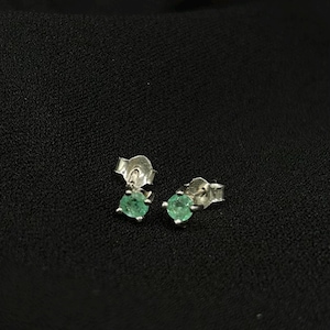 May include: A pair of stud earrings with small, round, light green gemstones set in silver. The earrings are displayed on a black surface, showcasing their simple elegance and color.