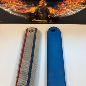 May include: Two rectangular gear straps with top holes. One strap is beige with vertical red and blue stripes. The other strap is solid blue with a textured surface. Behind them is a rectangular sign featuring a firefighter with fire wings and the text No Greater Love.