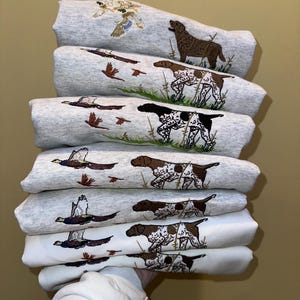 Op de afbeelding: Een stapel grijze sweatshirts met geborduurde ontwerpen met een jachtthema. Elk sweatshirt heeft een ander ontwerp, waaronder honden, vogels en gras. De sweatshirts zijn opgevouwen en op elkaar gestapeld.