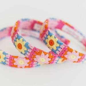 May include: Three colorful crochet headbands. Each headband features a floral pattern with pink, orange, yellow, and blue details. The headbands are arranged on a white surface.