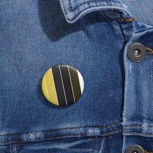 Puede incluir: Un botón circular con alfiler que presenta un patrón de rayas verticales en tonos amarillo, negro, verde oliva oscuro y blanco. El botón está sujeto a una tela de mezclilla azul con costuras visibles y un cierre de botón metálico oscuro.