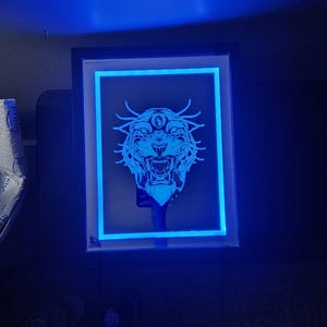 以下が含まれることがあります： 青い光を放つ、黒いフレームの長方形LEDライト。中央に詳細な、照らされた虎の頭のデザインがあり、青い縁取りがあります。アートワークは暗い背景に設定されています。