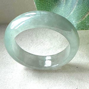 May include: A smooth, polished jade ring with a translucent, pale mint green colour. The band features a rounded, domed exterior surface and a circular opening, showcasing a natural, glossy finish that reflects light across its curved, solid stone structure.