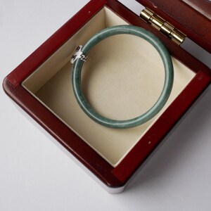 May include: A jade bangle bracelet is displayed in a wooden jewellery box. The bracelet is a pale green colour and appears translucent. The box is deep red with a light beige interior and gold-coloured hinges.