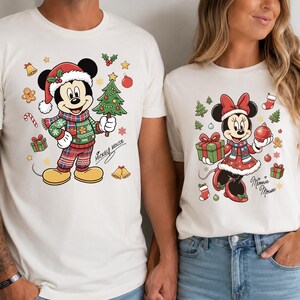 Puede incluir: Dos camisetas de color blanco roto con personajes de Disney con temática navideña. La camiseta de la izquierda muestra a Mickey Mouse con gorro de Papá Noel y suéter sosteniendo un árbol. La derecha muestra a Minnie Mouse con vestido festivo.