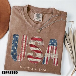 May include: A brown short-sleeved t-shirt with a crew neckline and a Comfort Colors brand tag. The front features the letters USA in a distressed, frayed fabric texture with a red, white, and blue flag pattern, and the text VINTAGE 1776 below.