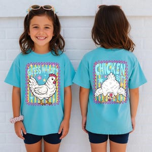 May include: A turquoise short-sleeve t-shirt featuring a graphic print of a white chicken. The front displays the chicken with the text "GUESS WHAT?" and the back shows the chicken from behind with the text "CHICKEN BUTT" over a colorful striped background.