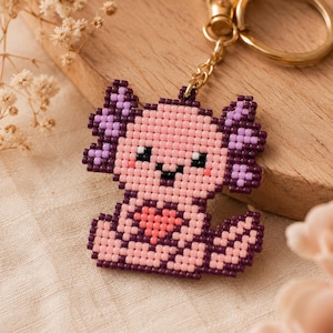 Könnte beinhalten: Ein Perlen-Schlüsselanhänger in Form eines Axolotls im Pixel-Art-Stil. Der Axolotl ist hellrosa mit dunkelvioletten Kiemen, schwarzen Augen und einem kleinen korallenfarbenen Herz auf der Brust. Er ist an einem goldfarbenen Ring befestigt.