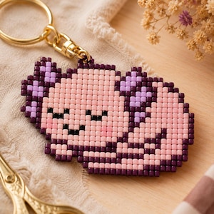 Peut inclure: Un porte-clés en perles représentant un axolotl de style pixel-art dans des tons rose clair, violet foncé et lavande. L'axolotl a les yeux fermés et une petite bouche en perles noires, fixé à un anneau brisé et un mousqueton dorés.