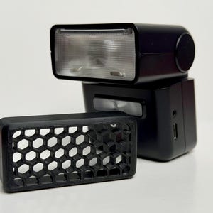 May include: A black camera flash unit with a rectangular head and a clear lens, accompanied by a separate black honeycomb grid attachment. The flash features a side port and a small silver rectangular light panel on the front of the main body.