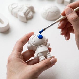 May include: A person is painting a small white astronaut figurine with a blue dot on the helmet. Other white figurines, including a planet and letters, are in the background. The astronaut is being painted with a small paintbrush.