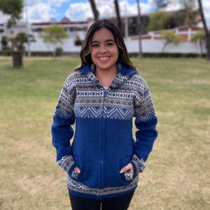 May include: A blue zip-up sweater with a patterned design in white, gray, and blue. The sweater has a hood and pockets. The person is wearing black pants. The background is a grassy area with trees and a building.