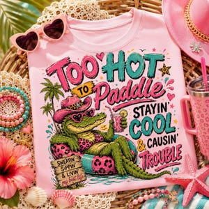 Peut inclure: T-shirt rose avec un dessin d'alligator portant un chapeau et des lunettes de soleil, se prélassant sur un anneau gonflable. Le texte dit "Too Hot to Paddle, Stayin' Cool & Causin' Trouble". Des lunettes de soleil en forme de cœur et un chapeau rose sont également visibles.