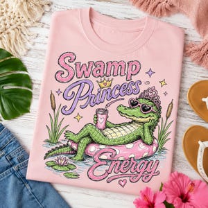 Könnte beinhalten: Hellrosa T-Shirt mit einer Grafik eines Alligators mit Sonnenbrille und Krone, der auf einem rosa Schwimmring liegt. Der Text lautet "Swamp Princess Energy" in einer verspielten Schriftart. Das Design umfasst Sterne, Wasserpflanzen und ein Getränk.